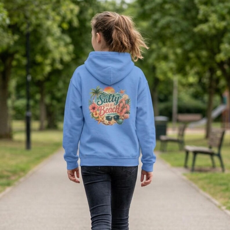 May include: A light blue hooded sweatshirt featuring a graphic on the back with the text Salty at a Beach, surrounded by illustrations of a palm tree, hibiscus flower, starfish, sunglasses, and flip-flops, worn with dark denim jeans.
