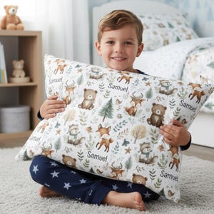 Op de afbeelding: Een witte kussensloop met een bosdierenpatroon, waaronder beren, vossen en herten, met de naam "Samuel" erop gedrukt. De kussensloop maakt deel uit van een beddengoedset. De jongen draagt een blauwe pyjama met witte sterren.
