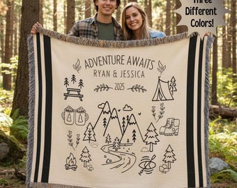 May include: A cream-colored woven throw blanket featuring black line art of mountains, trees, a tent, a campfire, and mugs. The text reads ADVENTURE AWAITS, RYAN & JESSICA, and 2025. The blanket has black striped borders and fringed edges.
