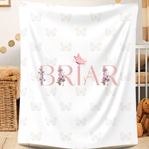 May include: White baby blanket with a repeating butterfly pattern. The name "BRIAR" is displayed in pink, with floral accents and a pink butterfly. The blanket is made of soft material, perfect for a nursery.