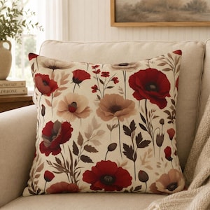 May include: A square throw cushion featuring a botanical print on a cream-coloured background. The design displays multiple red and beige poppy flowers with dark centres, accompanied by thin stems, dark brown leaves, and small buds scattered throughout.