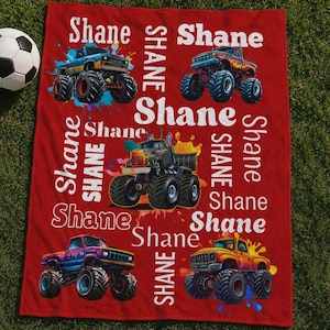 May include: Red blanket with multiple monster truck illustrations and the name "Shane" in white. The trucks are in various colours, with paint splatter accents. A football is visible in the upper left corner.