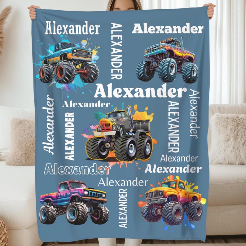 Op de afbeelding: Een blauwe deken met illustraties van kleurrijke monstertrucks en de naam "Alexander" in het wit. De trucks zijn afgebeeld met grote banden en splash art. De naam "Alexander" wordt verticaal en horizontaal herhaald.
