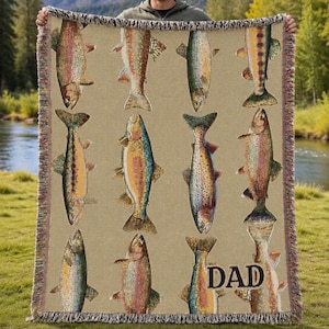 Puede incluir: Una manta tejida beige con un patrón repetido de ilustraciones de peces coloridos. Los peces son en tonos de naranja, verde y azul. La palabra "DAD" está impresa en negro en la parte inferior derecha. La manta tiene un borde con flecos.