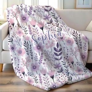 May include: A white floral blanket with a pattern of pink and purple flowers and foliage. The blanket is draped over a white sofa. The name "Sophia" is written in cursive across the blanket.