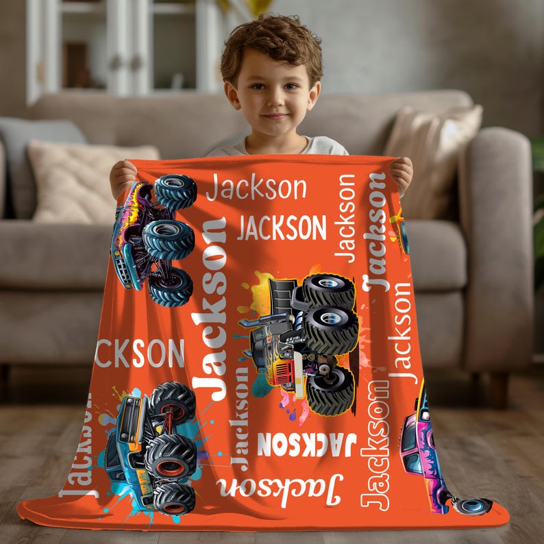 Op de afbeelding: Een oranje deken met kleurrijke monstertruckafbeeldingen en de naam "Jackson" in het wit. De deken heeft een levendig ontwerp met kleurspatten en wordt vastgehouden door een jong kind.