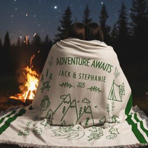 May include: A beige camping blanket with green detailing and a nature-themed design. The blanket has the text "ADVENTURE AWAITS" and personalised details. The design includes mountains, trees, a tent, and other camping-related illustrations.