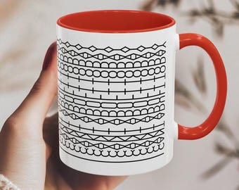 Puede incluir: Una taza de cerámica blanca con un interior rojo vibrante y un asa roja a juego. La superficie exterior está decorada con un patrón gráfico negro repetitivo que consiste en bandas horizontales de formas geométricas, incluyendo rombos y círculos.