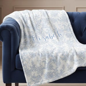 May include: A white and blue floral patterned blanket draped over a navy blue sofa. The blanket features a delicate floral design and the name "Elizabeth" in a light blue script.