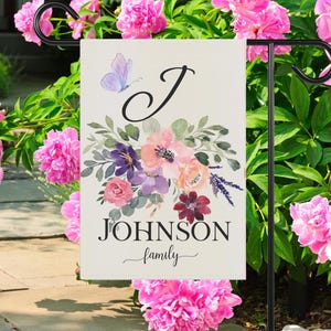 May include: A white garden flag featuring a watercolour floral design in shades of pink, purple, and burgundy. The letter "J" is in a cursive font and the text "JOHNSON family" is below the flowers.