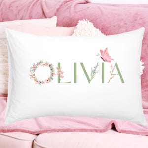 May include: White pillowcase with the name "OLIVIA" in green letters. The letter "O" is a floral wreath, and a pink butterfly is next to the letter "A". The pillowcase is on a pink couch.