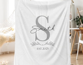 Könnte beinhalten: Eine weiße Decke mit einem großen grauen Buchstaben "S" und dem Namen "Smith" in Schreibschrift. Darunter ein dekorativer Zweig und der Text "EST 2025". Die Decke ist drapiert und zeigt ihre Textur.