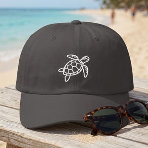 May include: A charcoal grey baseball cap featuring a white embroidered sea turtle graphic on the front panel. The cap has a curved brim and a structured crown. A pair of brown tortoiseshell sunglasses rests on the brim of the baseball cap.