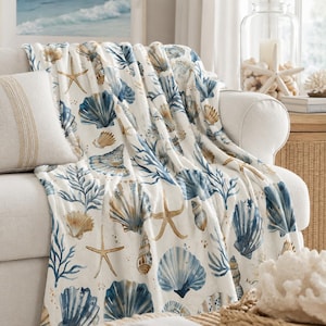 May include: A soft throw blanket featuring a repeating pattern of various seashells, starfish, and coral branches in shades of blue, tan, and white. The design is printed on a cream-colored fabric, showcasing detailed illustrations of marine life.
