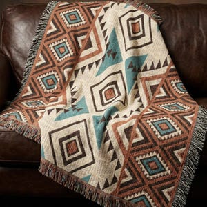May include: A woven throw blanket featuring a geometric pattern of diamonds and triangles in shades of cream, brown, and teal. The blanket has a textured surface and is finished with a fringed edge along the sides, showcasing a repeating design.