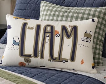 Könnte beinhalten: Ein rechteckiges Dekokissen mit cremefarbenem Hintergrund und dem Namen LIAM in großen dunkelblauen Buchstaben. Das Design zeigt kleine Illustrationen von Baufahrzeugen, Bäumen und Wolken in Orange-, Blau- und Grüntönen.