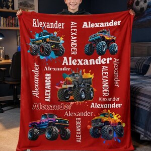 Op de afbeelding: Rode fleece deken met kleurrijke monster truck illustraties en de naam "Alexander" in het wit. De deken heeft meerdere monster truck ontwerpen met verfspatter effecten, waardoor het een leuk en persoonlijk cadeau is.