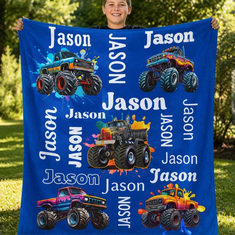 Op de afbeelding: Een blauwe deken met illustraties van kleurrijke monstertrucks en de naam "Jason" in het wit. De trucks zijn in verschillende kleuren, met grote banden en splash art achtergronden. De deken wordt door een persoon vastgehouden.
