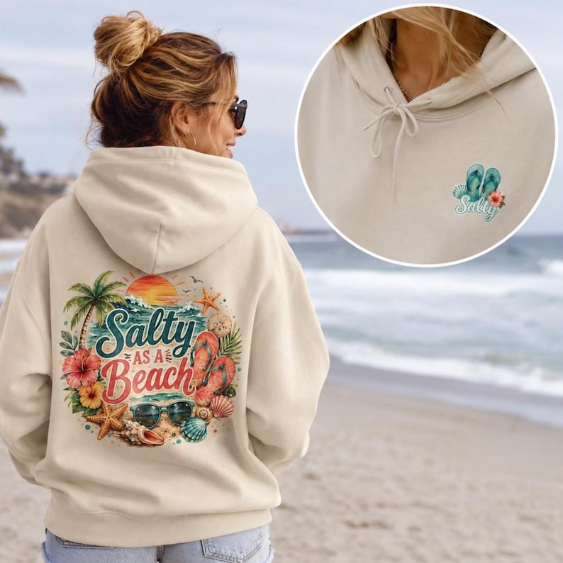 May include: A beige hooded sweatshirt featuring a large back graphic of a beach scene with a palm tree, starfish, seashells, flip-flops, sunglasses, and the text Salty as a Beach. The front chest area includes a small matching flip-flop graphic.