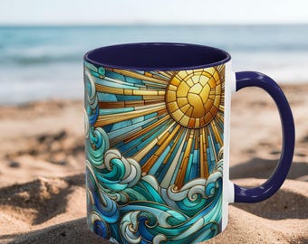 May include: White ceramic mug with a navy blue interior and handle. The mug features a stained-glass design of a golden sun with blue and teal waves. The mug is sitting on a sandy surface near the sea.