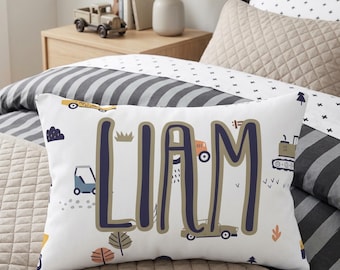 Könnte beinhalten: Ein rechteckiges weißes Dekokissen mit dem Namen LIAM in großen, dunkelblauen Buchstaben mit beiger Umrandung. Das Kissen ist mit einem Muster aus verschiedenen Baufahrzeugen bedruckt, darunter ein gelbes Auto, ein blauer LKW und ein grüner Traktor.