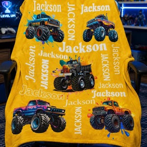 Op de afbeelding: Mosterdgele deken met kleurrijke monster truck ontwerpen en de naam "Jackson" in het wit. De trucks zijn in verschillende kleuren, waaronder blauw, paars en oranje, met grote banden en spat effecten.