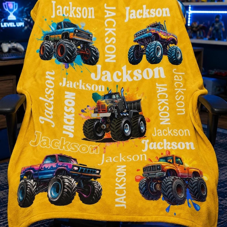 Op de afbeelding: Mosterdgele deken met kleurrijke monster truck ontwerpen en de naam "Jackson" in het wit. De trucks zijn in verschillende kleuren, waaronder blauw, paars en oranje, met grote banden en spat effecten.