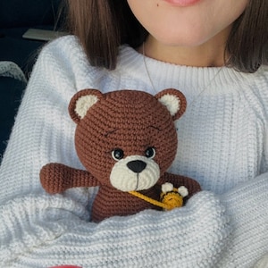 May include: A brown crocheted teddy bear with a yellow crocheted bee charm. The bear is being held by a person wearing a white sweater.