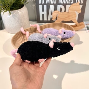 Puede incluir: Tres peluches de rata de crochet con orejas y narices rosas. Las ratas están en una cesta y una está siendo sostenida en una mano. La rata en la mano es negra, la rata en la cesta de la derecha es morada y la rata en la cesta de la izquierda es gris.