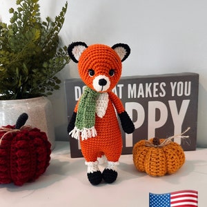 May include: A crocheted fox toy wearing a green scarf. The fox is orange with black eyes and a white nose. The toy is standing on a white surface with a sign that says "What makes you happy" and two crocheted pumpkins.