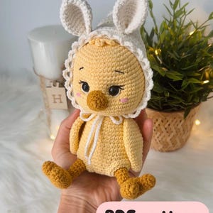 May include: A crocheted yellow chick toy wearing a white crocheted bunny hat with floppy ears. The toy is sitting in a person's hand. The image includes the text "PDF pattern ENG".