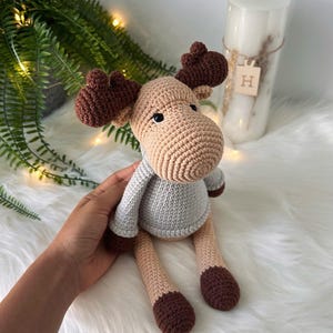 Puede incluir: Un juguete de alce de crochet que lleva un suéter azul claro. El alce tiene cuernos marrones, una nariz marrón y ojos negros.