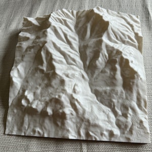 May include: A white 3D printed model of a mountain range. The model is detailed with contour lines and shows the peaks and valleys of the mountains.