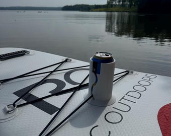 May include: A white stand-up paddleboard with black text reading ROC OUTDOORS, featuring black bungee cords secured by metal D-rings and a white cylindrical beverage holder containing a blue and silver aluminum can on the deck surface.