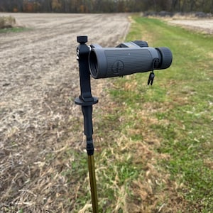 May include: A black and gray monocular mounted on a hiking stick, set against a backdrop of a field and trees. The monocular has a textured grip and a lanyard. The hiking stick is gold and black.