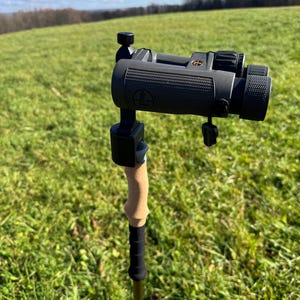 May include: A black and gray binoculars mounted on a brown and black hiking stick. The binoculars have a textured surface and a black strap. The hiking stick has a cork grip and a black rubber grip.