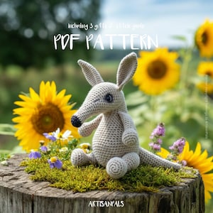 Può includere: Un giocattolo di oritteropo all'uncinetto è seduto su un ceppo muschioso, circondato da girasoli e fiori selvatici. L'oritteropo è grigio chiaro con naso e occhi neri. L'immagine include il testo "PDF PATTERN" e "Including 3 gifts & stitch guide."