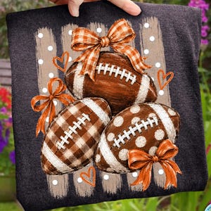 May include: A dark gray t-shirt featuring a graphic print of three brown footballs. The footballs are decorated with orange and white plaid bows, white polka dots, and white gingham patterns, accented by small orange heart outlines.