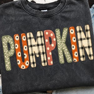May include: A charcoal gray acid-wash sweatshirt featuring the word PUMPKIN printed in large letters. Each letter displays a unique pattern, including green leaves, orange daisies, black and white plaid, white polka dots, and orange stripes.