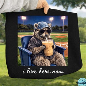 Può includere: Immagine quadrata nera con un procione con occhiali da sole e un berretto da baseball, seduto su una sedia blu, che beve da una tazza. Il testo "i live here now" è in basso. Lo sfondo mostra un campo da baseball.