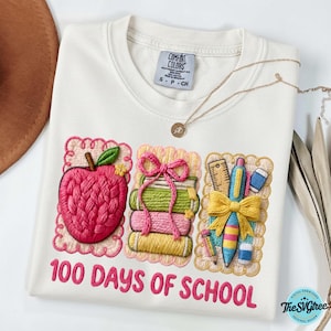 Pu&ograve; includere: Felpa Comfort Colors bianca con ricami a tema scolastico. Presenta una mela, libri e materiale scolastico. Il testo "100 DAYS OF SCHOOL" &egrave; ricamato in rosa. Una collana d'oro &egrave; drappeggiata sulla maglietta.