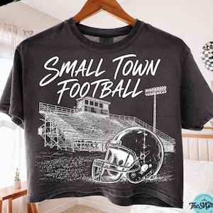 Puede incluir: Una camiseta gris carbón de manga corta con un estampado gráfico blanco de una grada de estadio de fútbol y un casco. El texto "SMALL TOWN FOOTBALL" está impreso en una fuente cursiva blanca sobre la ilustración del estadio y el casco.