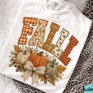 Op de afbeelding: Een wit t-shirt met het woord FALL in grote blokletters, elk met een ander herfstpatroon zoals oranje gingham, bloemen, ruiten en stippen. Onder de tekst staan pompoenen, bladeren en een beige strik in de stijl van jute.
