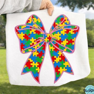 May include: White towel featuring a large bow design made of colorful puzzle pieces. The bow has a pink outline and the puzzle pieces are red, yellow, blue, and green. The towel is held up against a green background.