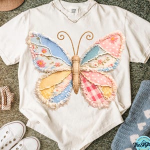 Può includere: T-shirt bianco con un design a farfalla patchwork colorato. Le ali della farfalla presentano motivi blu, rosa, gialli e floreali. La maglietta è realizzata in materiale morbido, adatta per un abbigliamento casual.