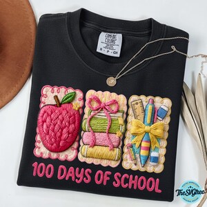Pu&ograve; includere: Felpa nera con toppe ricamate di una mela, libri e materiale scolastico. Il testo "100 DAYS OF SCHOOL" &egrave; ricamato in rosa. Una collana d'oro &egrave; drappeggiata sulla maglia.