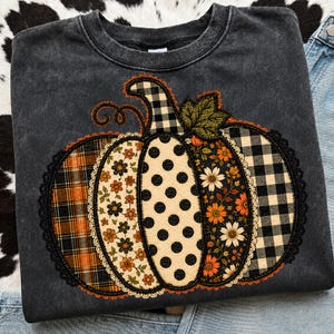 May include: A charcoal gray crewneck sweatshirt featuring a large central pumpkin graphic. The pumpkin is divided into five vertical segments with varied patterns including orange plaid, floral prints, black polka dots, and black and white buffalo check.