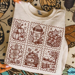 May include: A beige crewneck sweatshirt featuring a brown graphic print of six postage stamp-style illustrations. The designs include pumpkins, a coffee cup labelled FALL VIBES, a folded plaid blanket, a signpost, a truck, and a rural landscape.