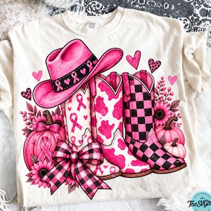 Puede incluir: Una camiseta de color crema con un estampado gráfico de un sombrero de vaquero rosa con lazos de concienciación sobre el cáncer de mama, dos botas vaqueras con estampado de vaca y cuadros rosas y negros, calabazas rosas, girasoles y corazones.