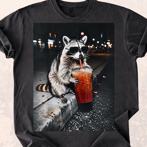 May include: Dark gray t-shirt featuring a raccoon wearing sunglasses and drinking from a clear plastic cup with a red straw. The raccoon is sitting on a concrete ledge with a blurred city background at night.